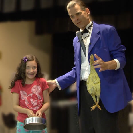 A young girl is laughing at a magician holding a rubber chicken during Chicago magic show!